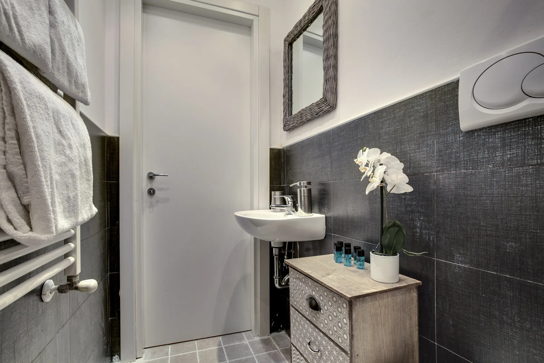 A bathroom with a wall-mounted sink, dark tiled walls, and a white door.