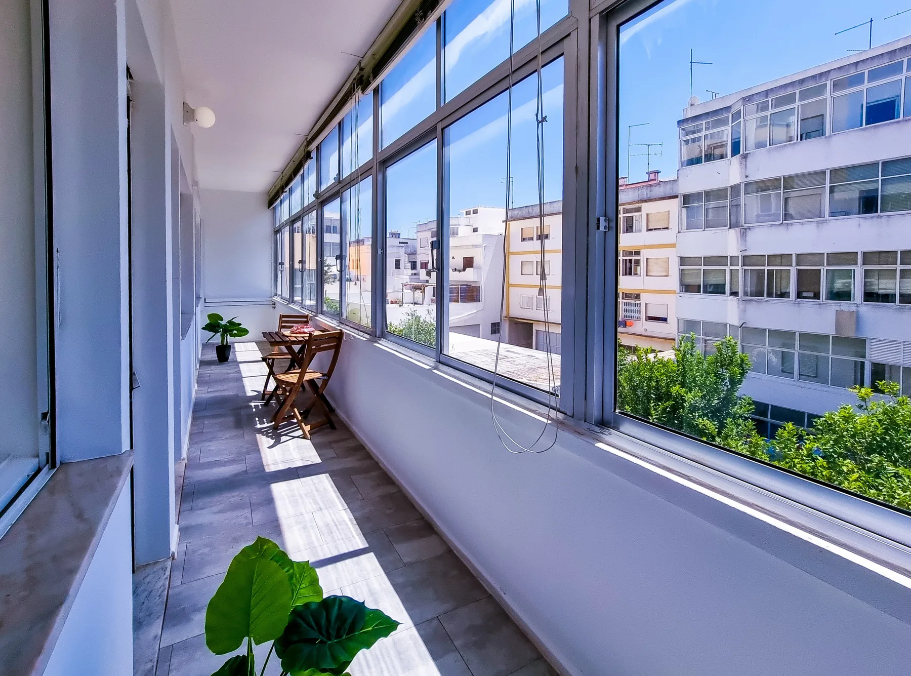 Un balcón cerrado y luminoso con grandes ventanas y una pequeña zona para sentarse.