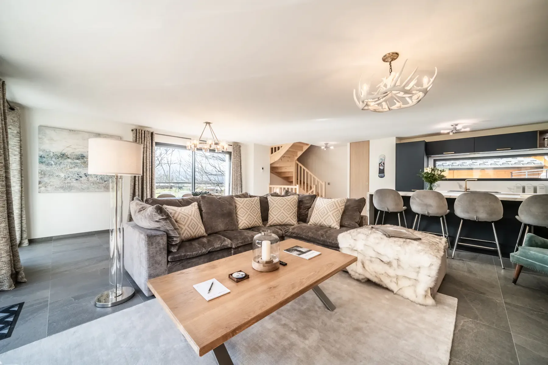 Spacious living room with a large gray sectional sofa, wooden coffee table, and contemporary chandelier. The room features large windows with views of greenery and a wooden staircase.