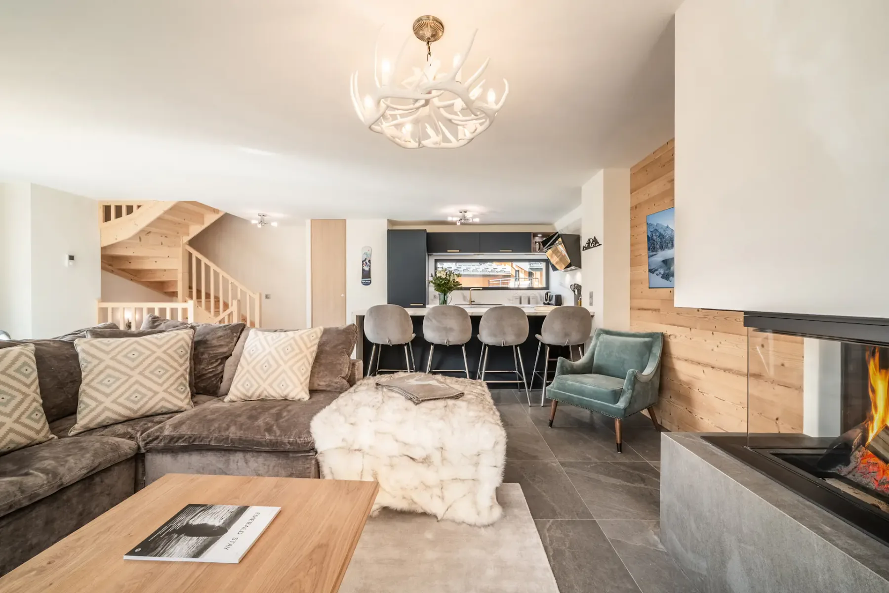 Spacious living room with a large gray sectional sofa, wooden coffee table, and contemporary chandelier. The room features large windows with views of greenery and a wooden staircase.