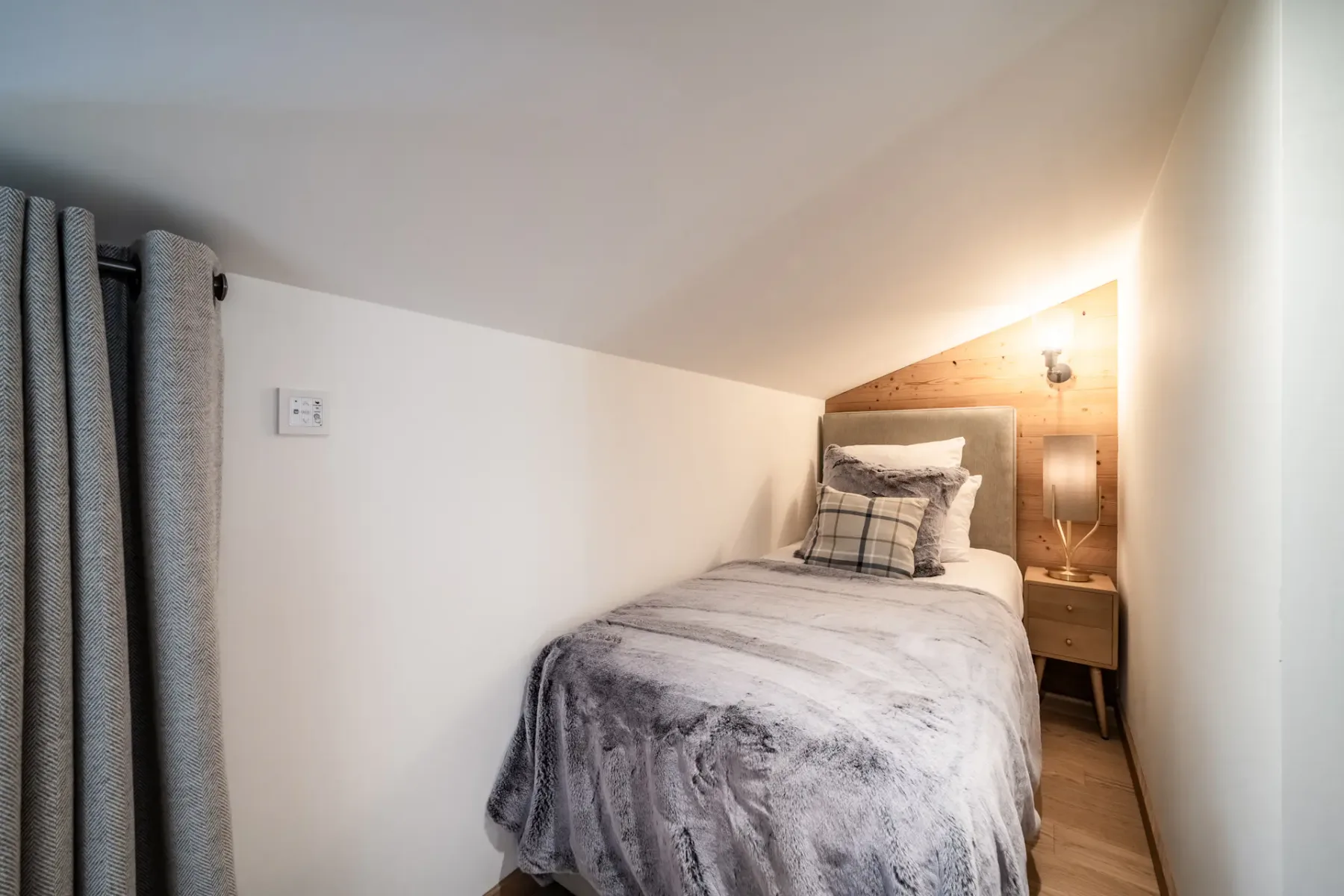 Cozy attic bedroom with a single bed, gray bedding, a plaid pillow, a wooden nightstand with a lamp, and a sloped ceiling.