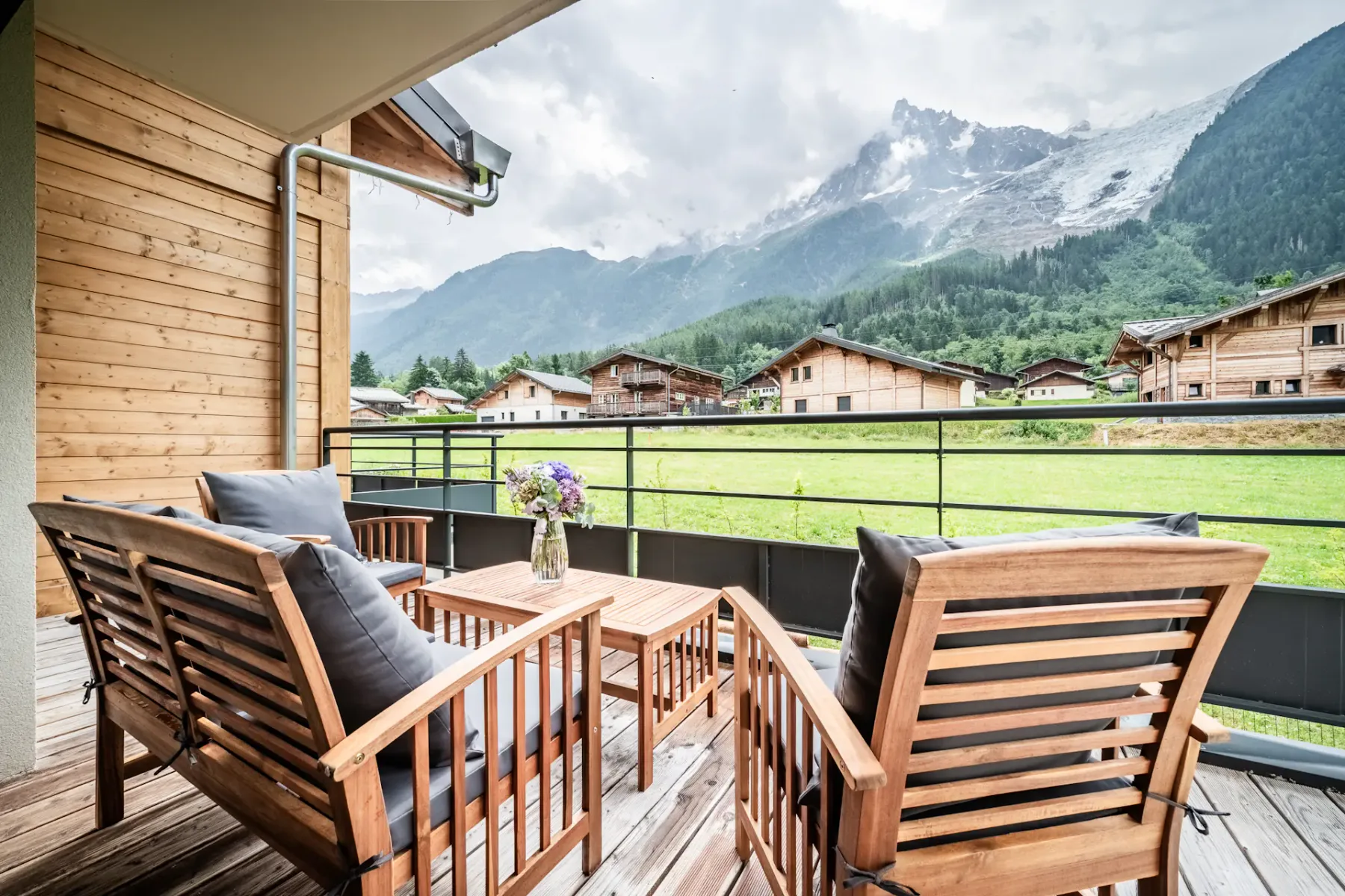 A wooden balcony with two chairs and a small table overlooks a grassy area, with sliding glass doors leading to a modern living room inside.