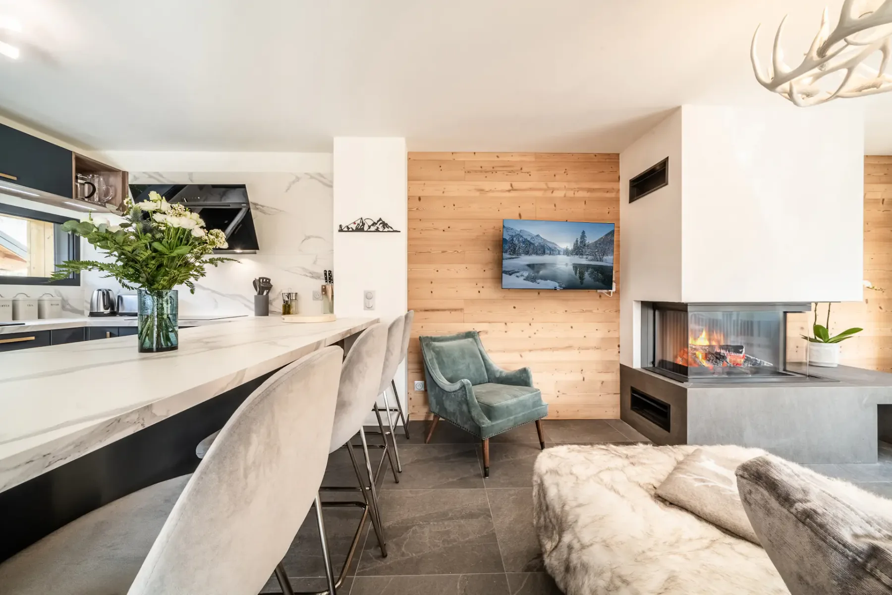 Spacious living room with a large gray sectional sofa, wooden coffee table, and contemporary chandelier. The room features large windows with views of greenery and a wooden staircase.