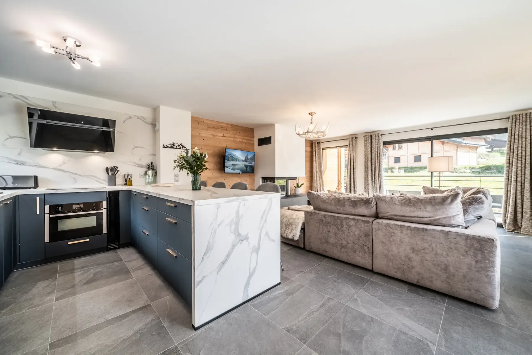 Modern open-plan living area with a kitchen featuring marble countertops, a spacious gray sofa, a dining table, a wall-mounted TV, and large windows overlooking a scenic view.
