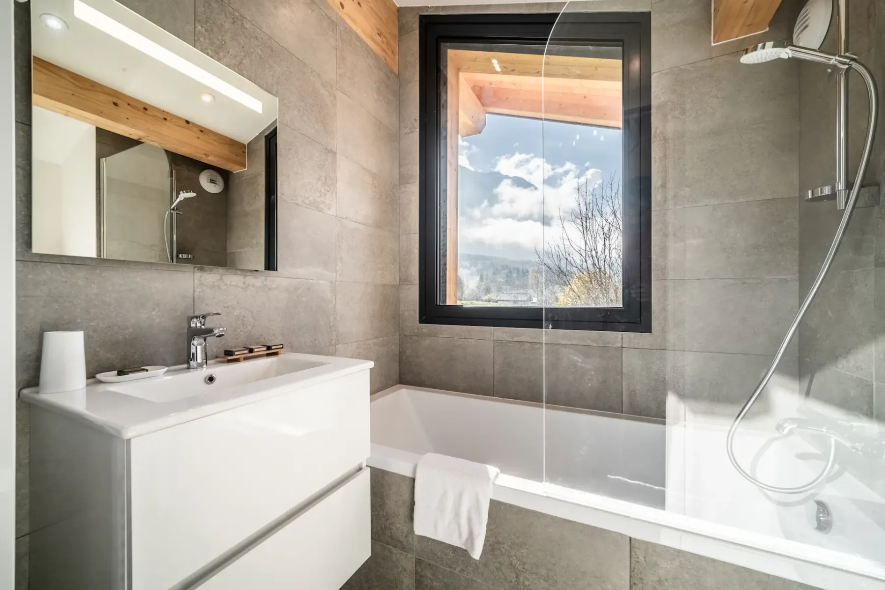 Modern bedroom and bathroom with wooden accents; the bathroom has a bathtub and a large window with a mountain view.