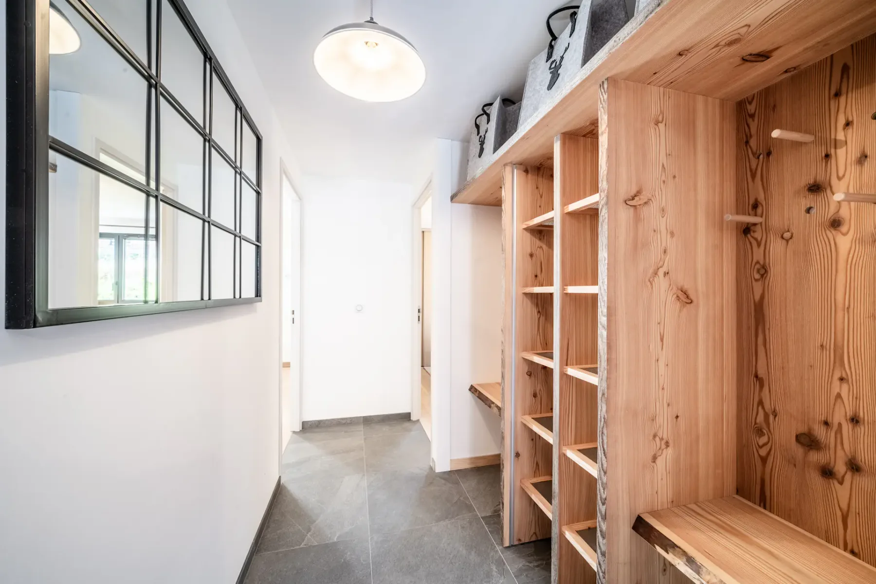 Wooden shelves and hooks line a hallway with gray tile flooring and a large black-framed mirror.