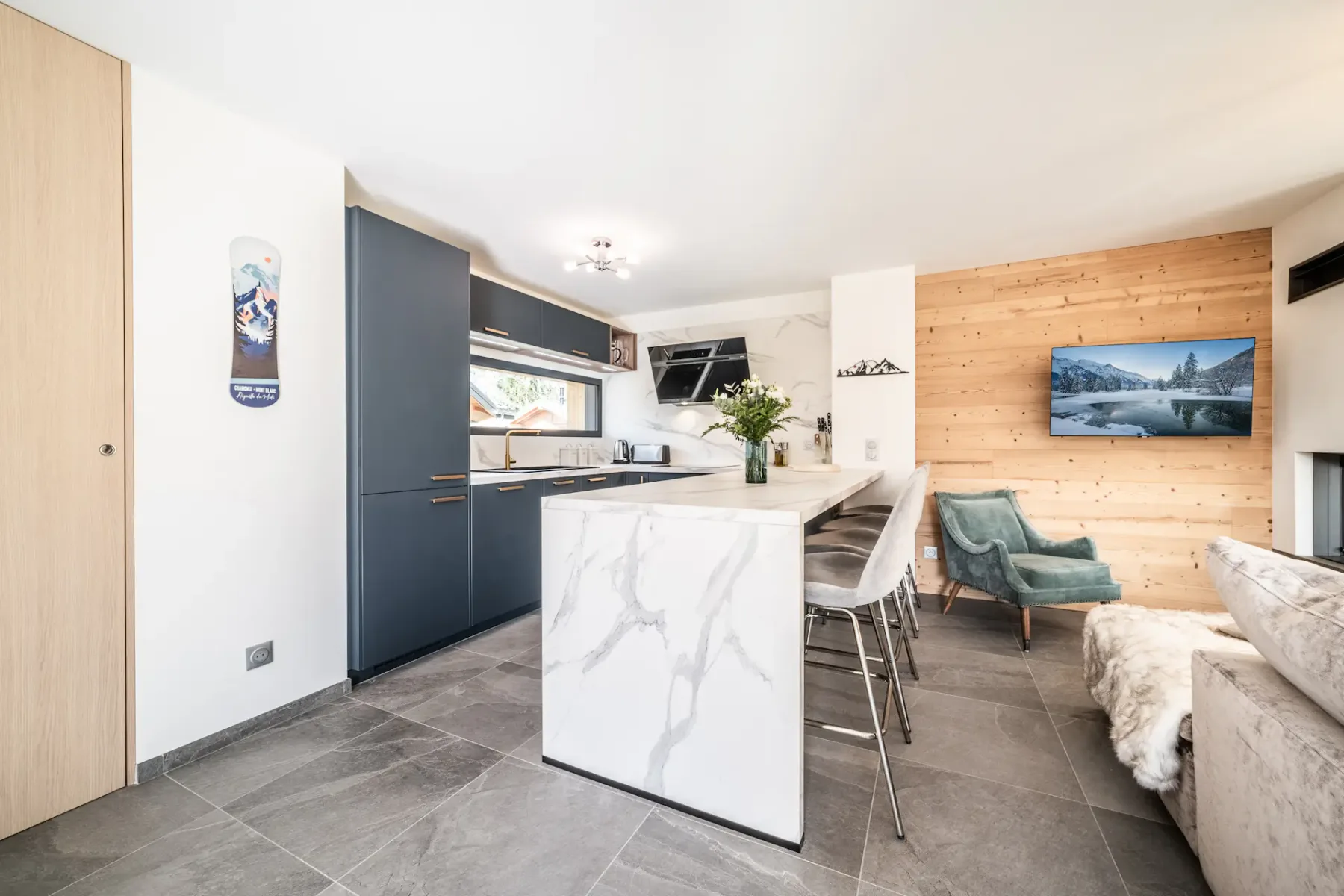 Modern open-plan living area with a kitchen featuring marble countertops, a spacious gray sofa, a dining table, a wall-mounted TV, and large windows overlooking a scenic view.