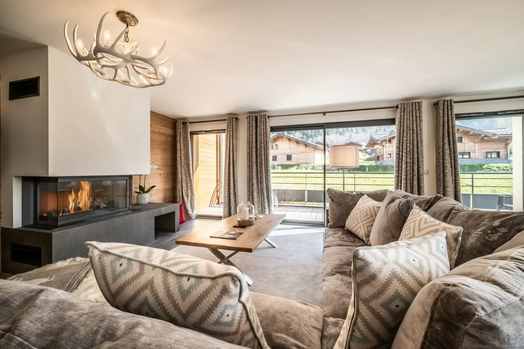Spacious living room with a large gray sectional sofa, wooden coffee table, and contemporary chandelier. The room features large windows with views of greenery and a wooden staircase.