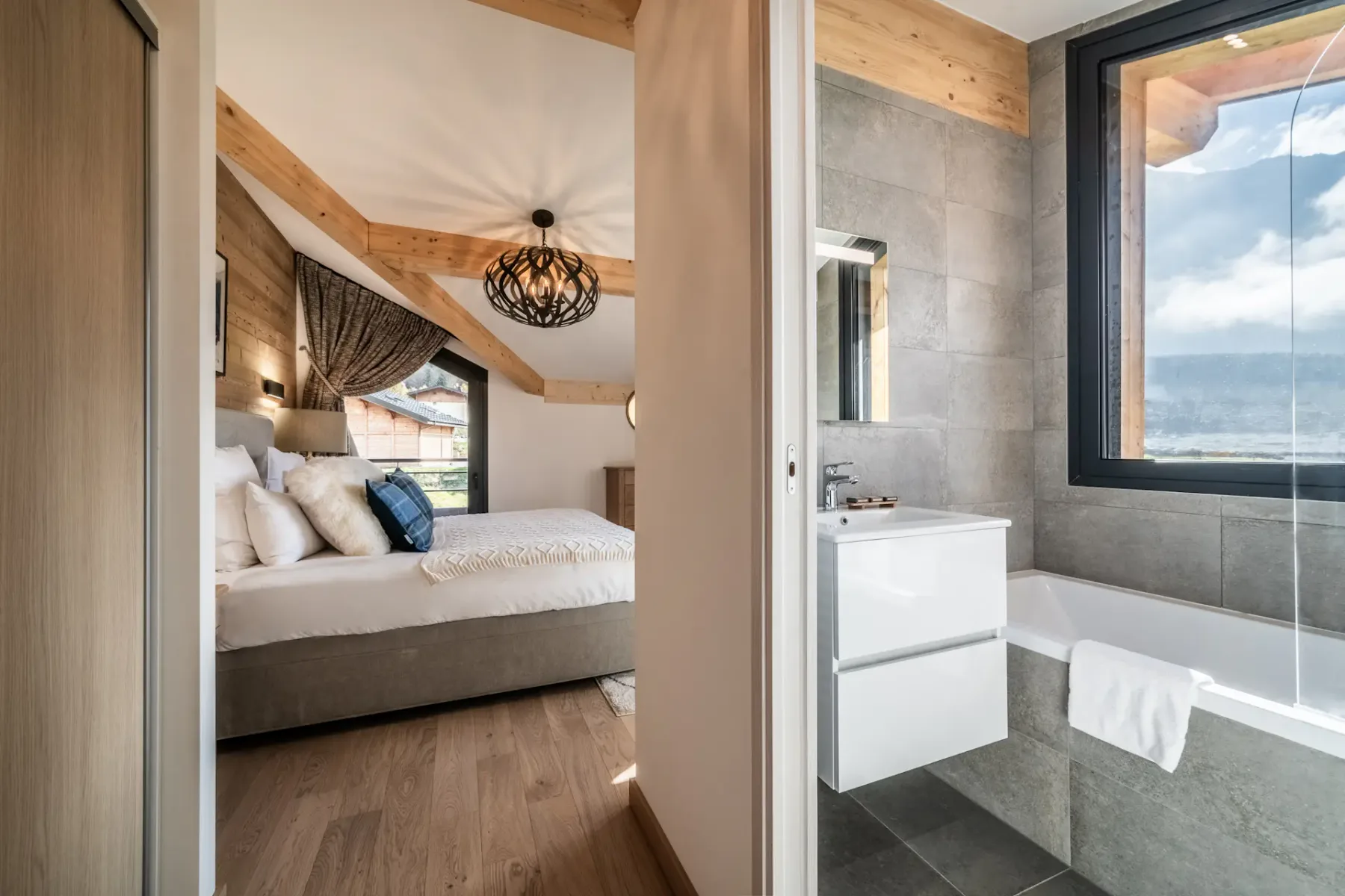 Modern bedroom and bathroom with wooden accents; the bathroom has a bathtub and a large window with a mountain view.