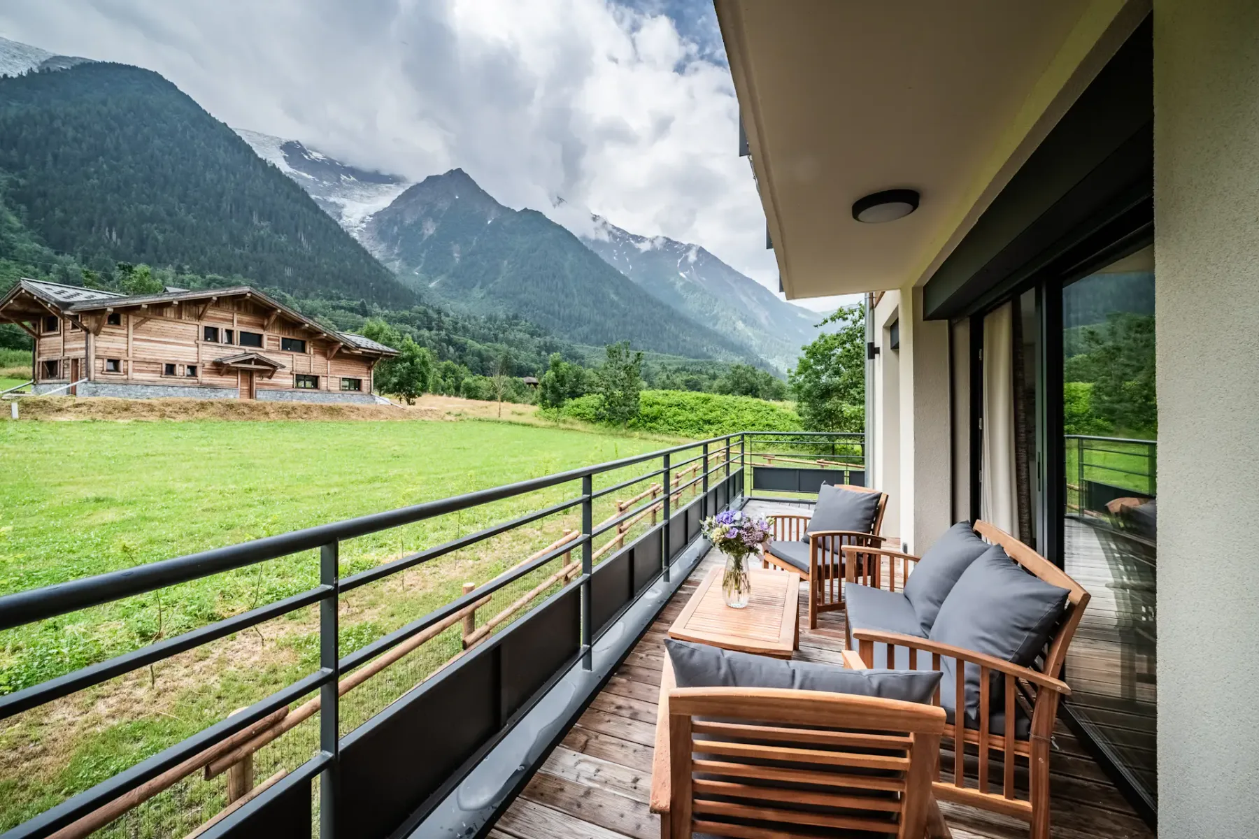 A wooden balcony with two chairs and a small table overlooks a grassy area, with sliding glass doors leading to a modern living room inside.