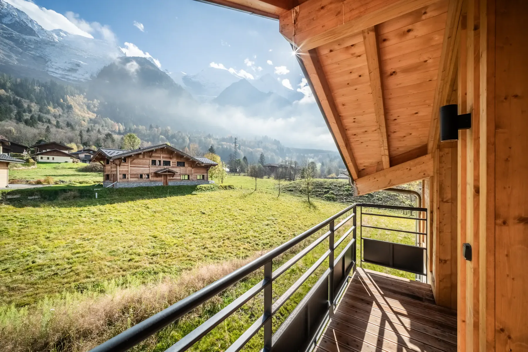 Scenic view from a wooden balcony overlooking a lush green field, distant chalet-style houses, and misty mountains under a bright sky.