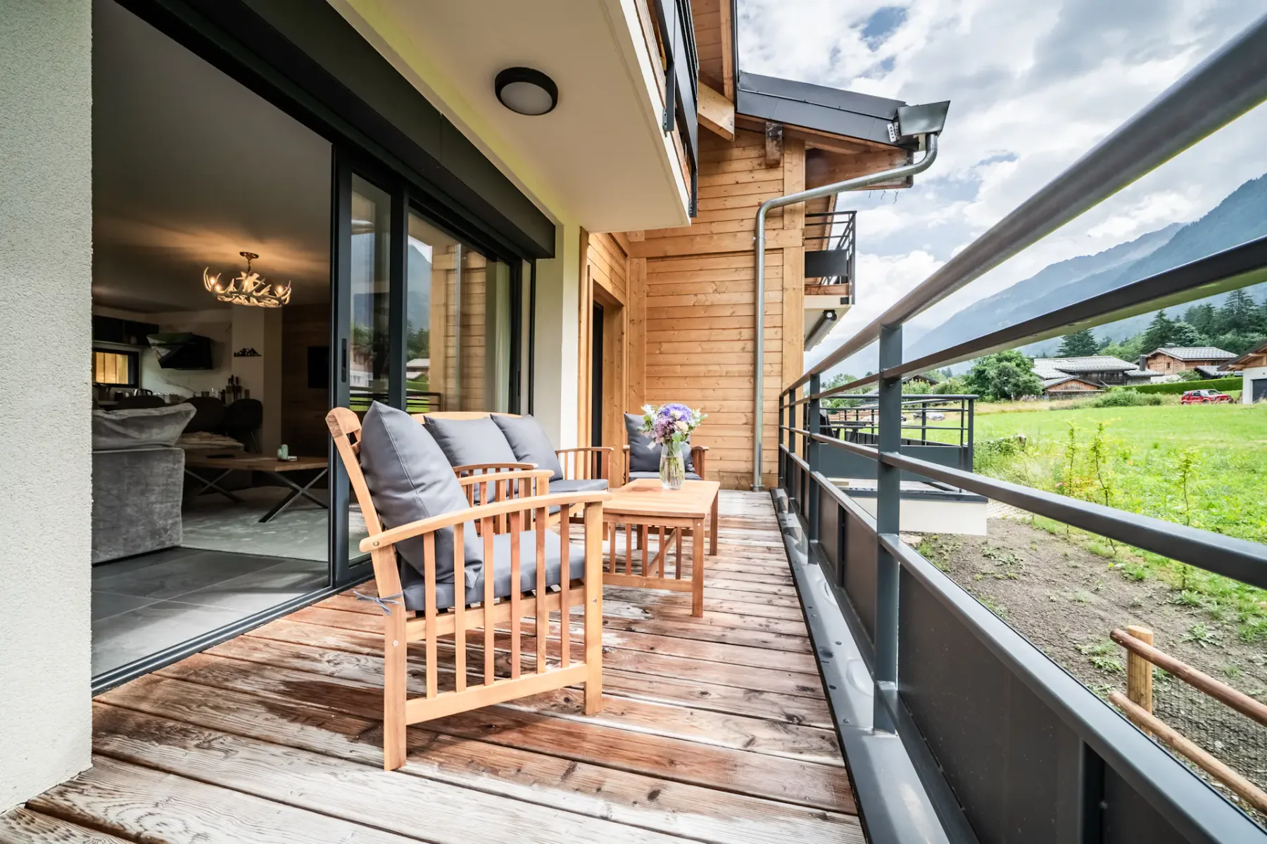 A wooden balcony with two chairs and a small table overlooks a grassy area, with sliding glass doors leading to a modern living room inside.