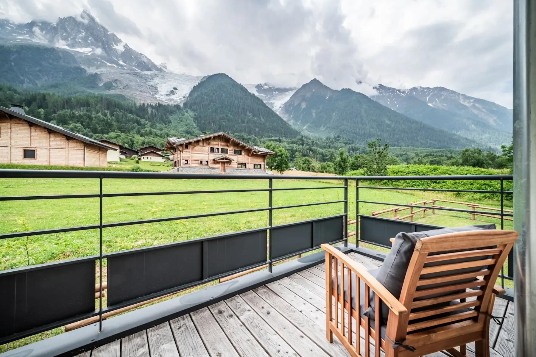 A wooden balcony with two chairs and a small table overlooks a grassy area, with sliding glass doors leading to a modern living room inside.