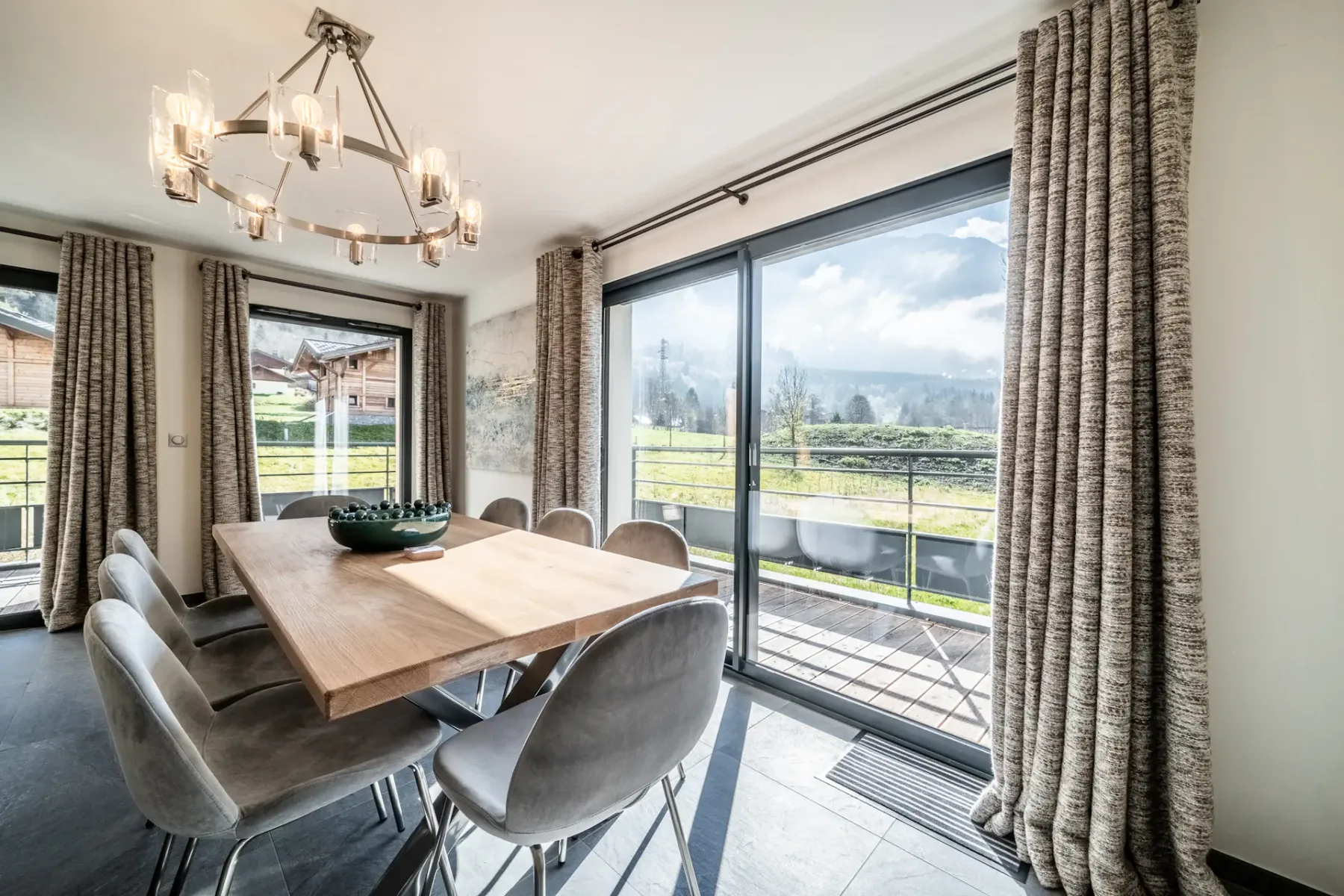 A dining room with a wooden table, six gray chairs, and a modern chandelier. Large windows with curtains offer a view of grass and hills outside.