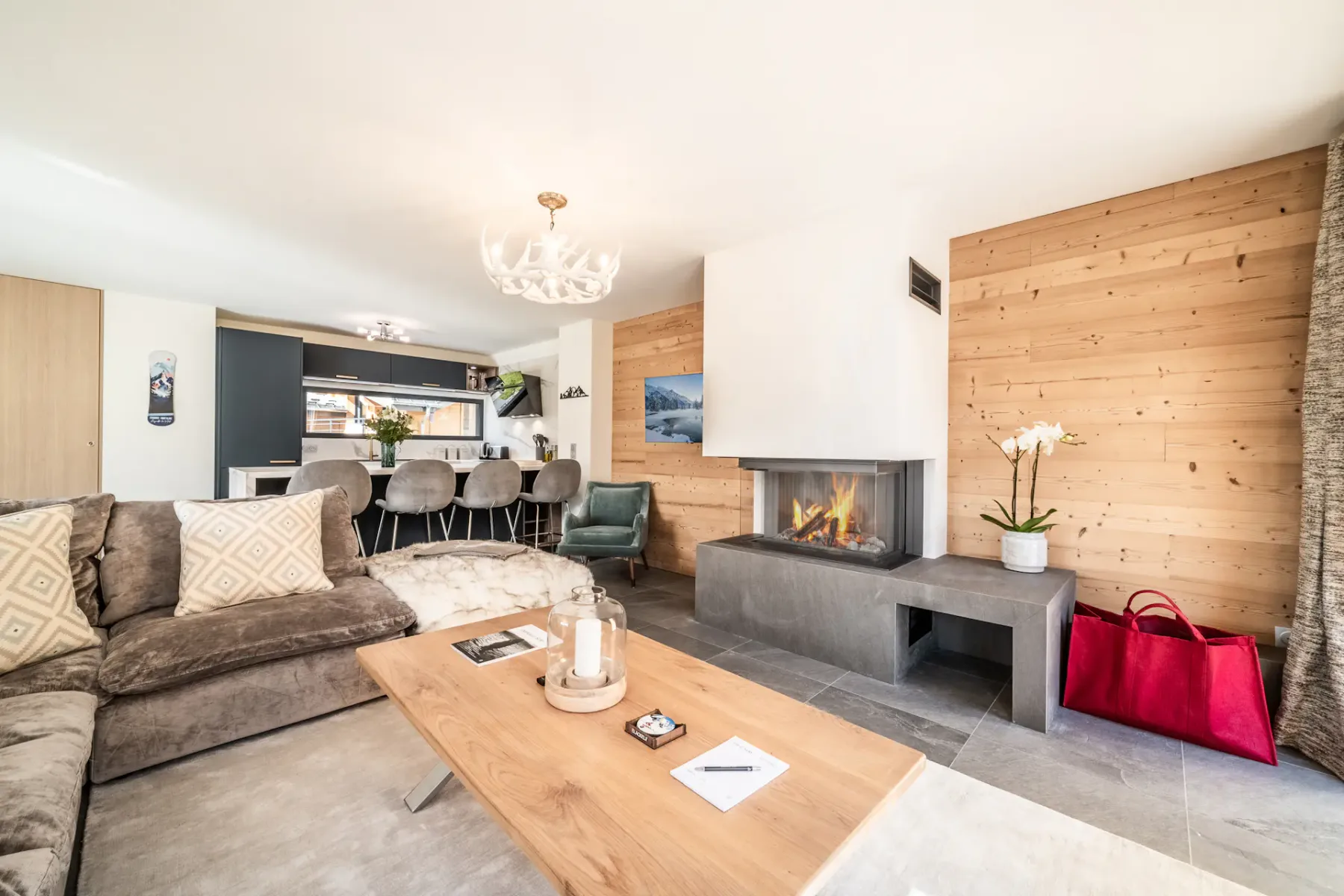 Spacious living room with a large gray sectional sofa, wooden coffee table, and contemporary chandelier. The room features large windows with views of greenery and a wooden staircase.