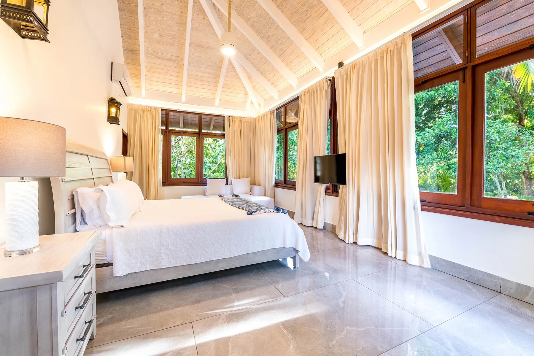 Un dormitorio luminoso con una cama grande, cortinas claras y ventanas con vistas a la vegetación.