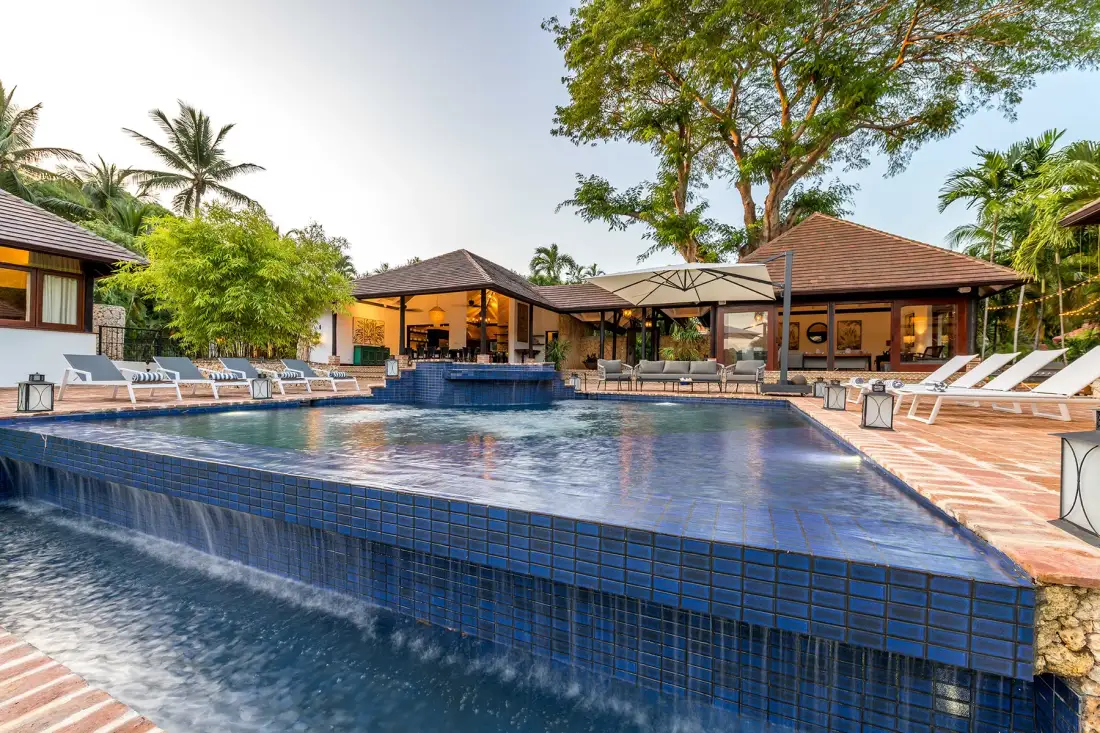 Una piscina junto a un patio con asientos y vegetación tropical.