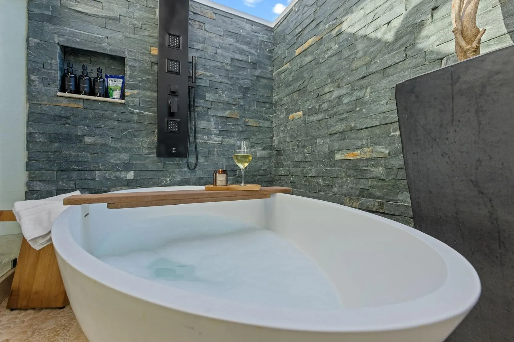 Una bañera blanca está en un baño con paredes de piedra y una ducha de pared.