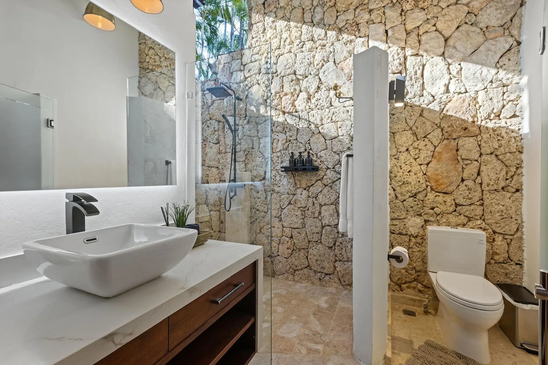 Un baño con lavabo, espejo, ducha e inodoro junto a una pared de piedra.