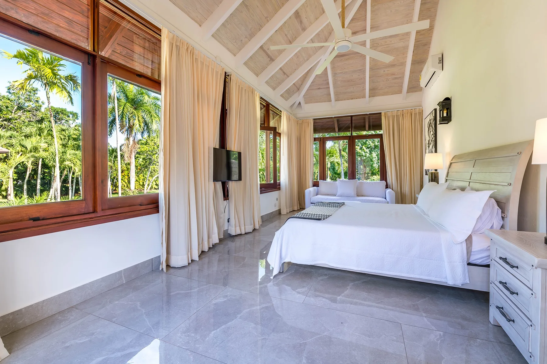 Un dormitorio luminoso con una cama grande, ventanas altas y un techo blanco con vigas.