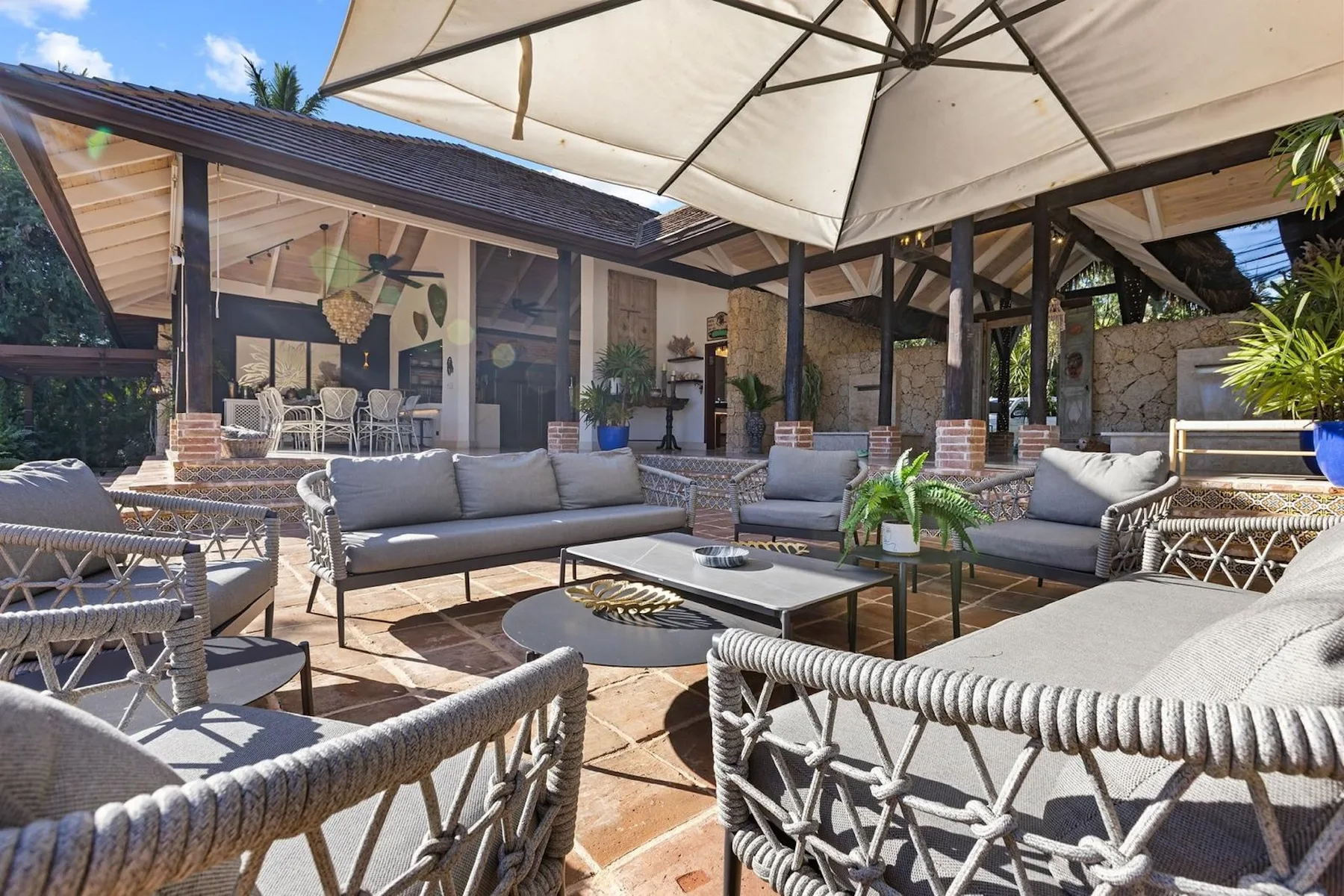 Un patio exterior cubierto con asientos alrededor de una mesa baja bajo una sombrilla grande.