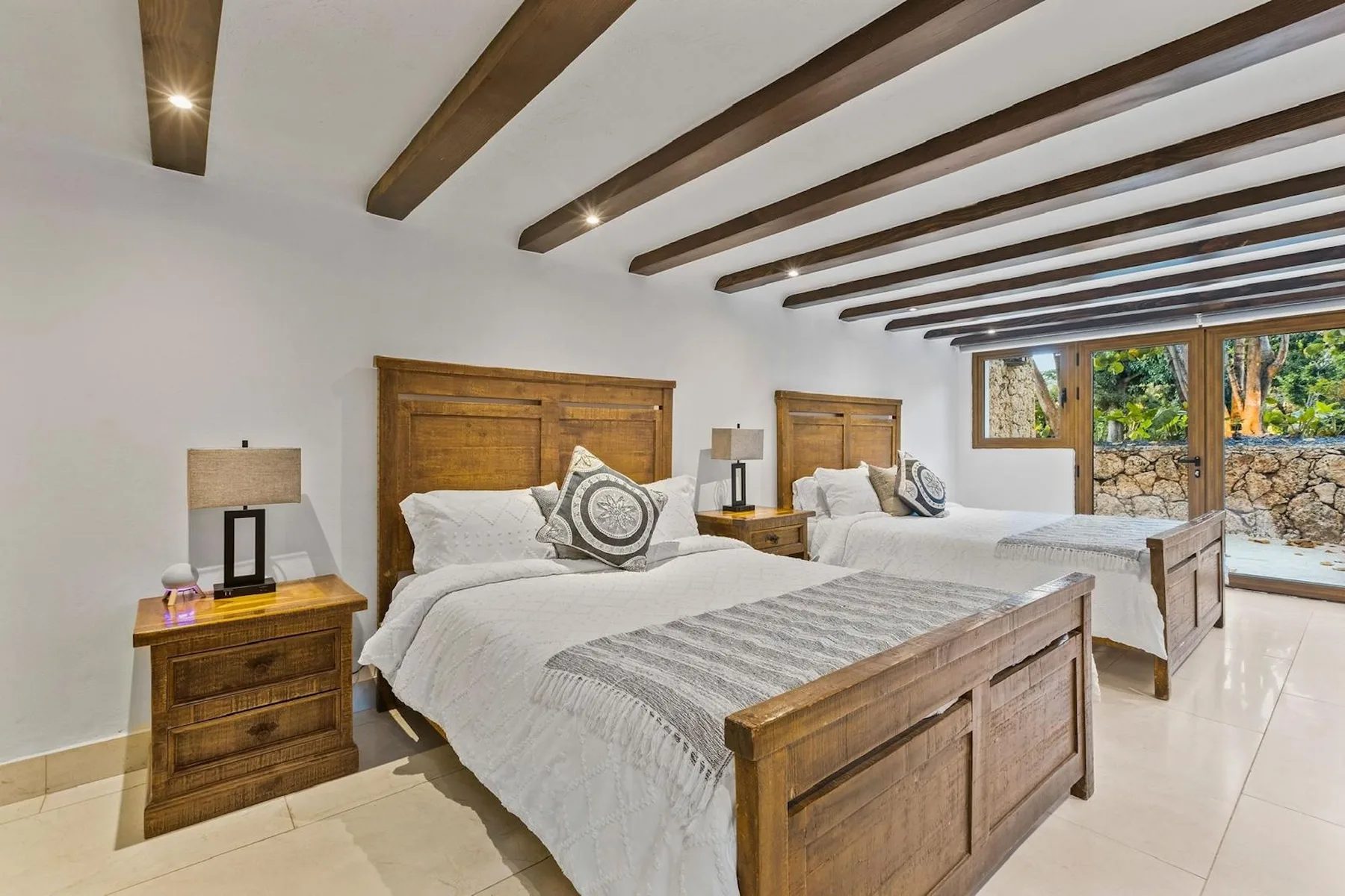 Un dormitorio luminoso con dos camas, cabeceros de madera y vigas vistas en el techo.