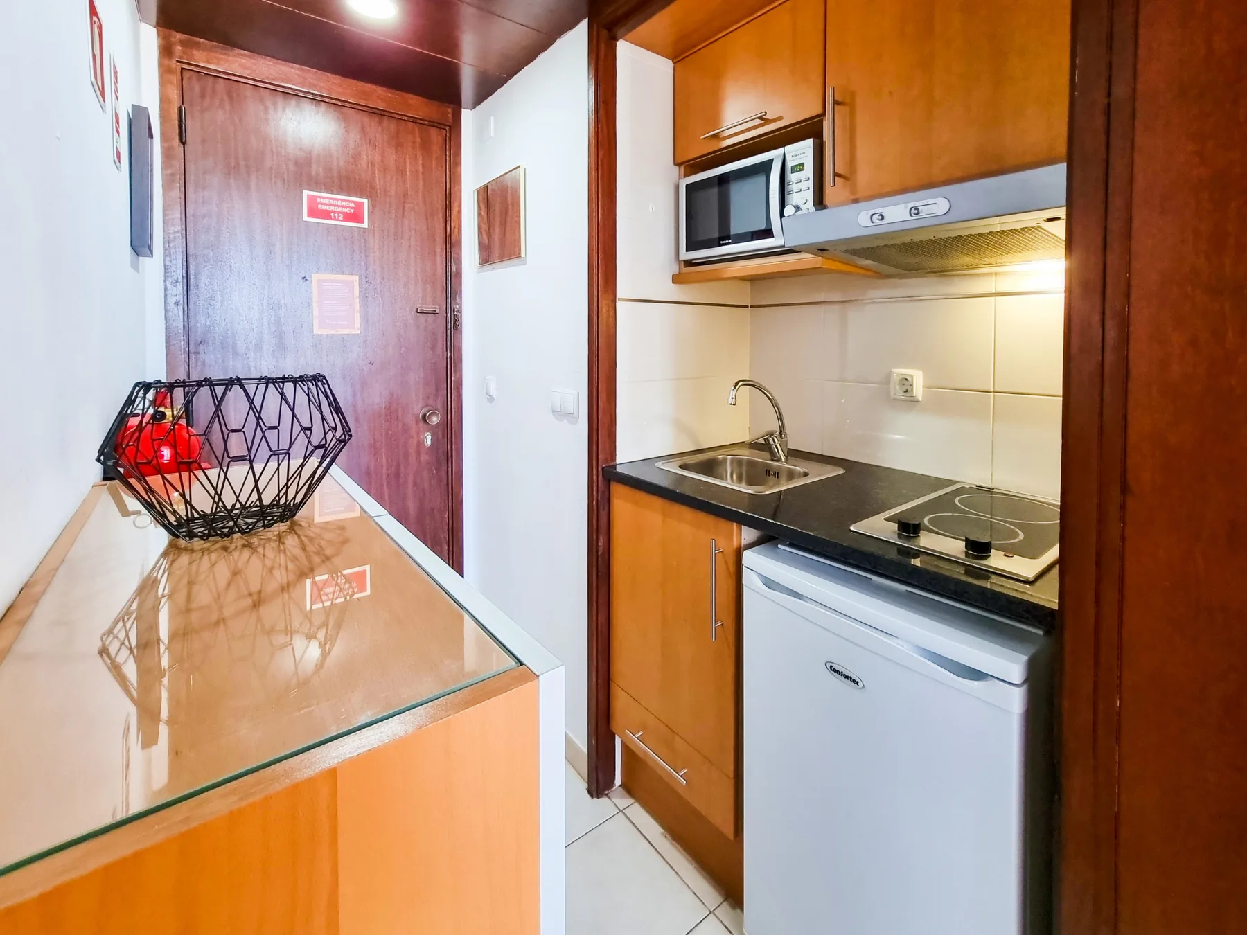 A compact kitchenette with a sink, cooktop, and microwave beside a glass-topped counter.