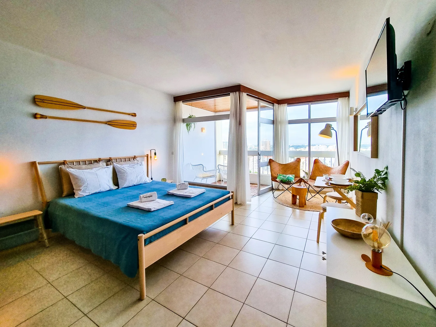 A bright studio room with a bed and a seating area beside large windows.