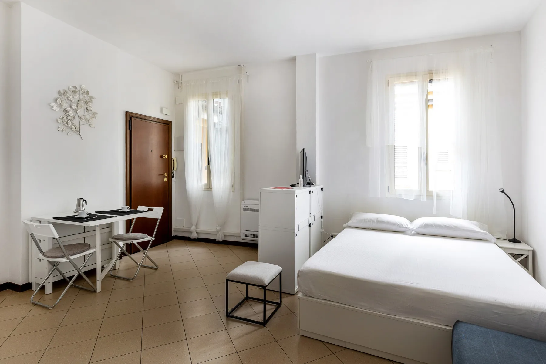 Un dormitorio luminoso con una cama, un escritorio pequeño y dos ventanas.