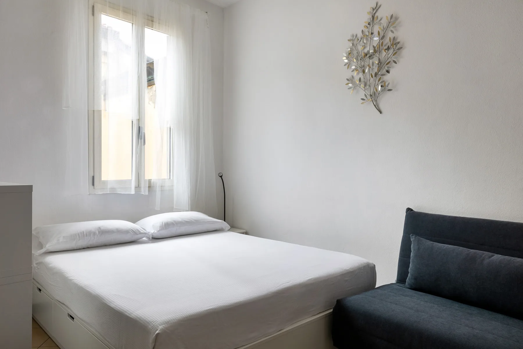 Un dormitorio luminoso con una cama junto a un asiento tapizado oscuro.