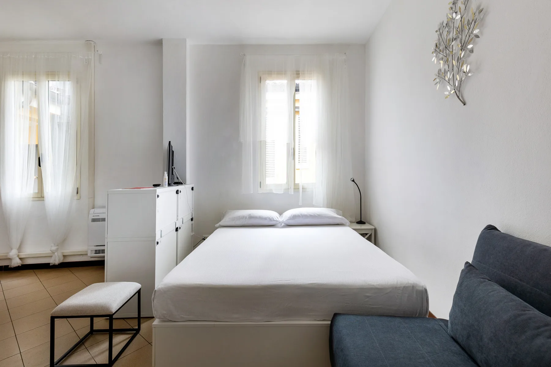 Un dormitorio luminoso con cama, ventanas con cortinas translúcidas y un sofá oscuro.