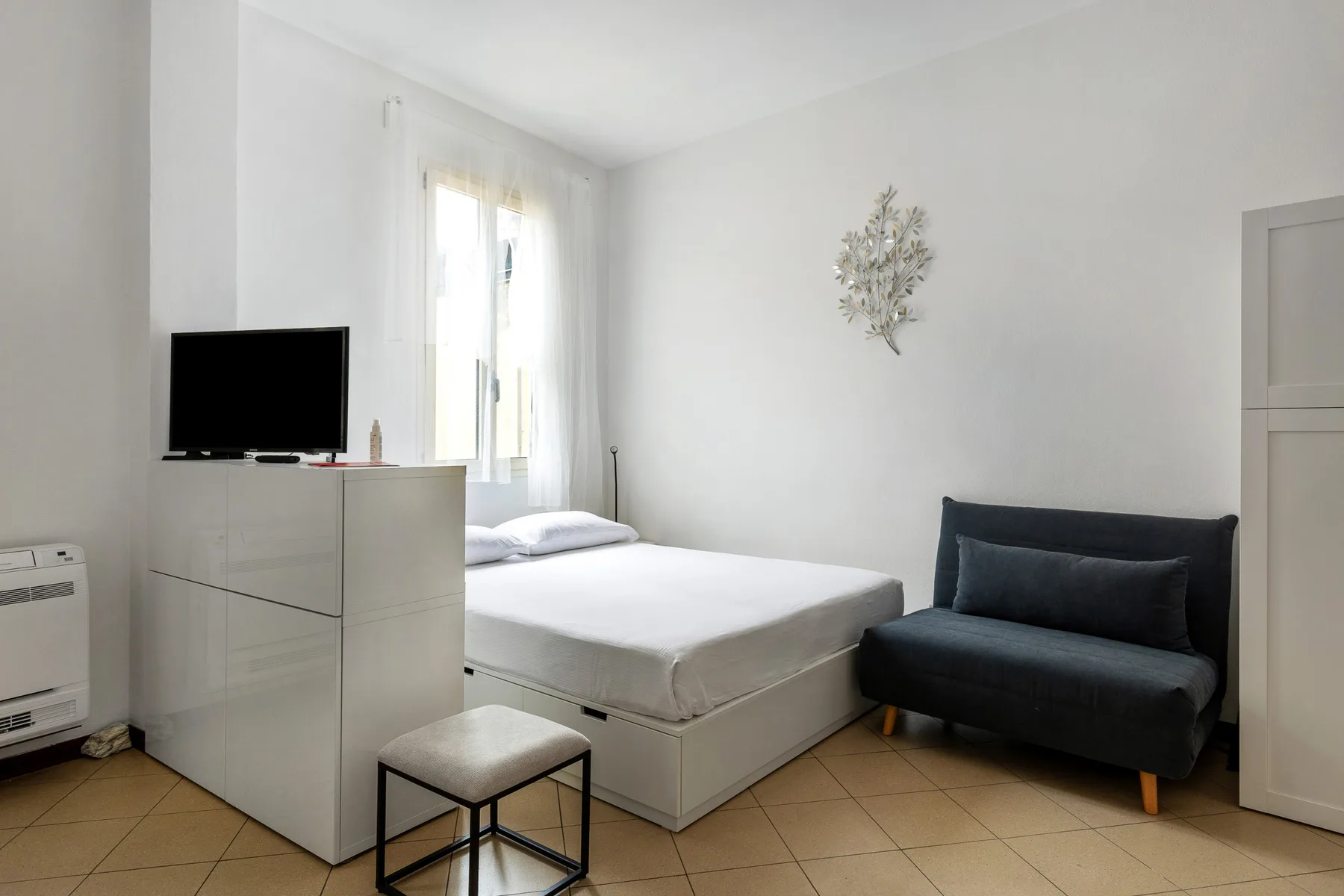 Un dormitorio luminoso con una cama, un sofá pequeño oscuro y un televisor sobre una cómoda.