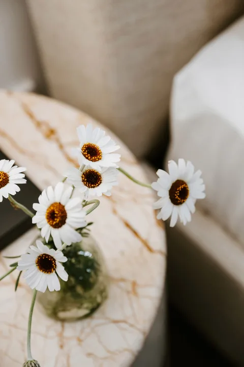 A small vase of white daisies sits on a round marble-patterned table beside a sofa.
