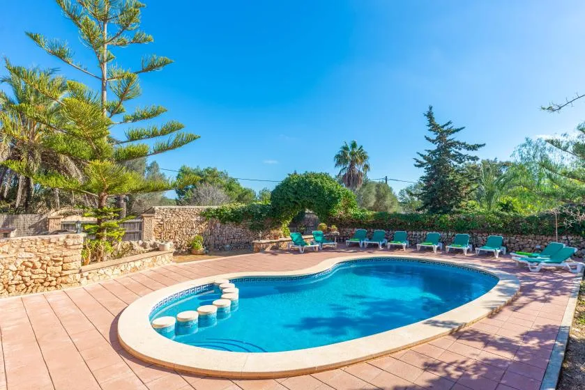 Piscina exterior rodeada por una terraza pavimentada, vegetación y un muro de piedra.