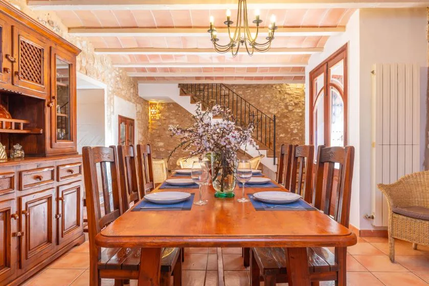 Una larga mesa de comedor está puesta con platos y un jarrón de flores bajo una lámpara de araña.