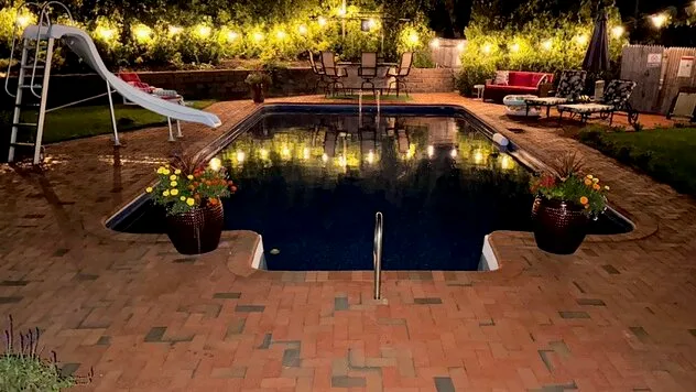 Una piscina exterior iluminada está rodeada de asientos de patio y luces colgantes.