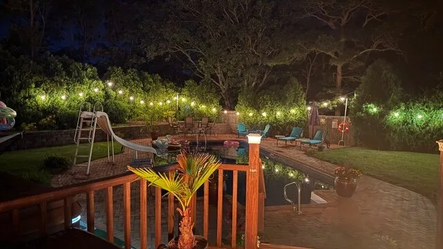 Una zona de piscina en un patio iluminada con calidez, con una terraza de madera y árboles alrededor.