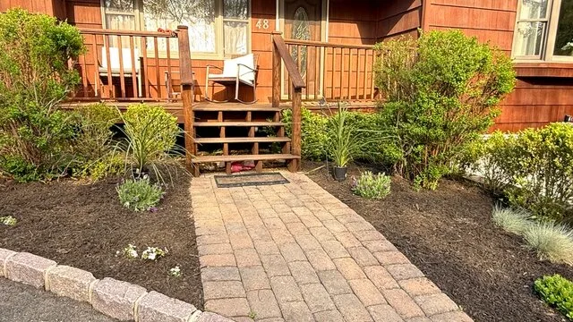 Un camino pavimentado conduce a un porche de madera junto a parterres.