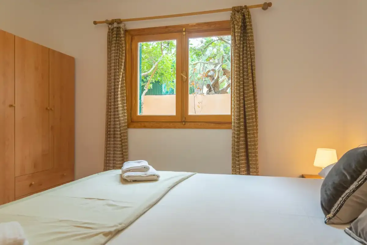 A bedroom with a neatly made bed, wooden wardrobe, and window overlooking greenery.