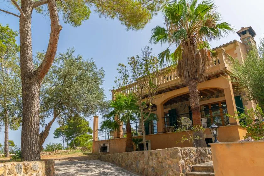 A tan building stands among trees and palms beside a stone walkway.