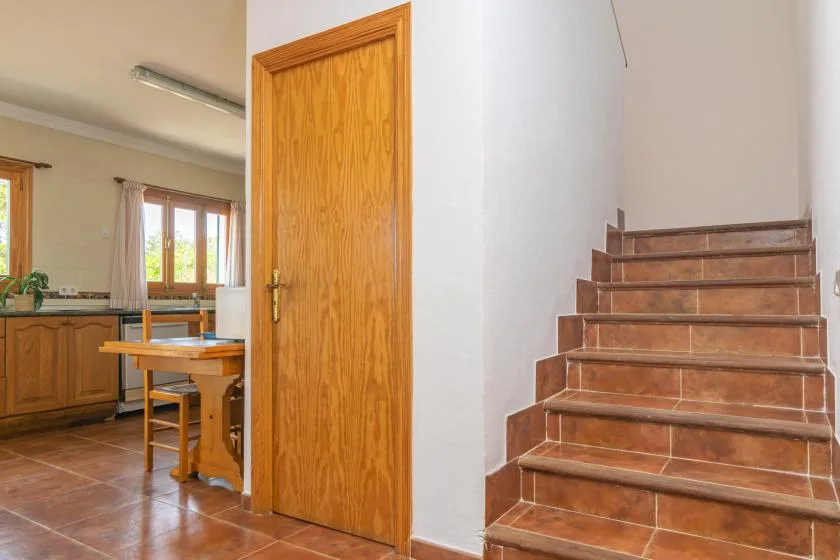 An interior staircase beside an open wooden door, with a kitchen visible beyond.