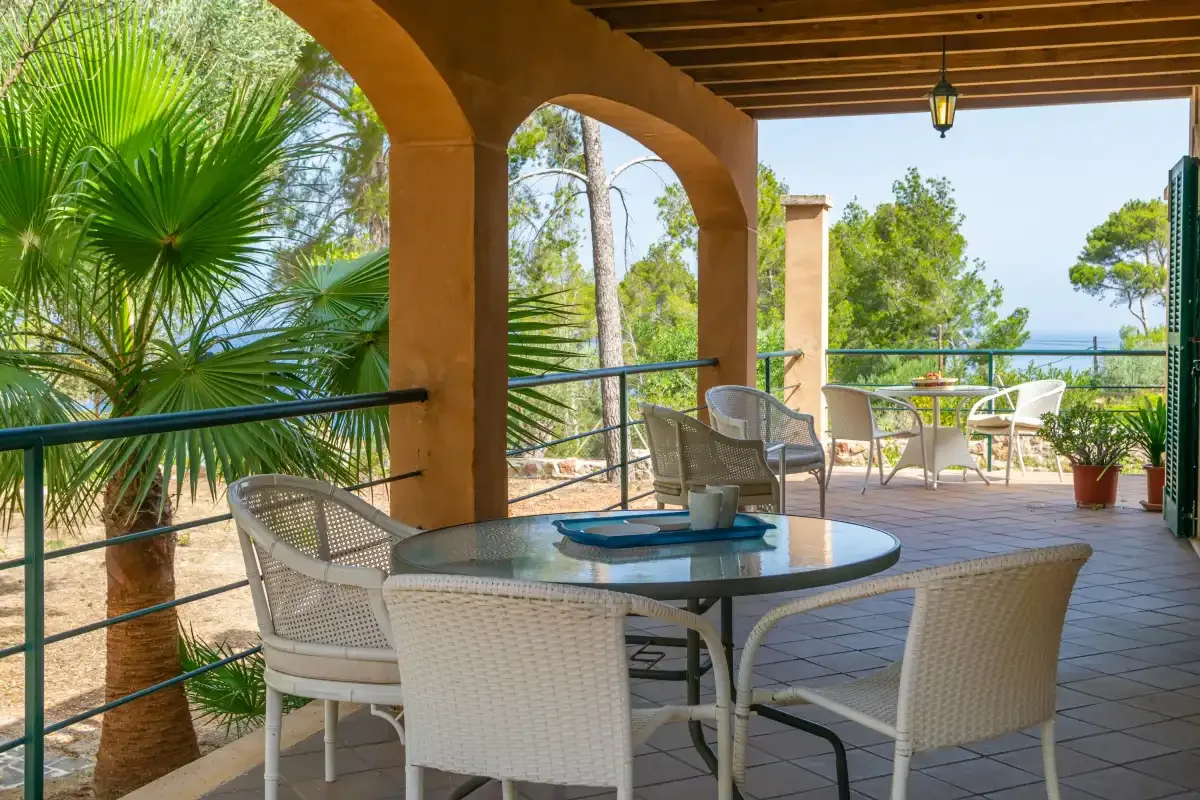 A covered terrace with a round table and seating overlooks greenery and the sea.