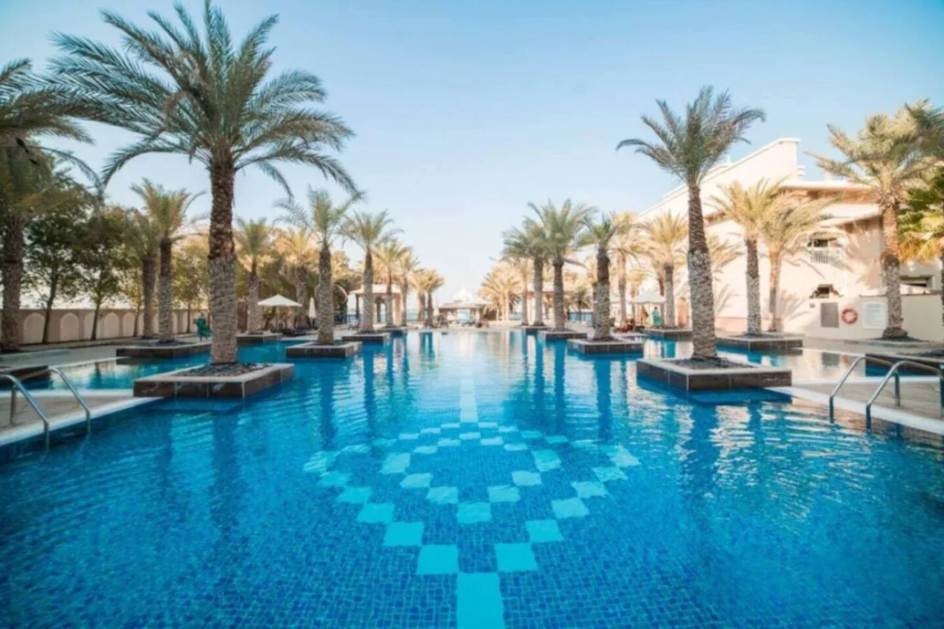 A large outdoor swimming pool is surrounded by palm trees and resort buildings.