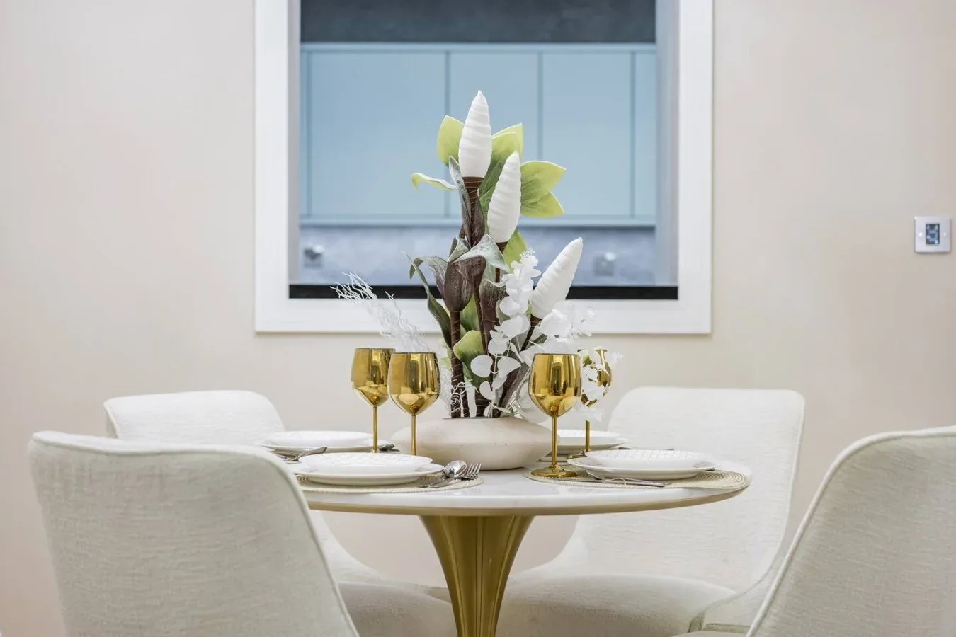 A dining area with a round table, pale seating, and a floral centerpiece.