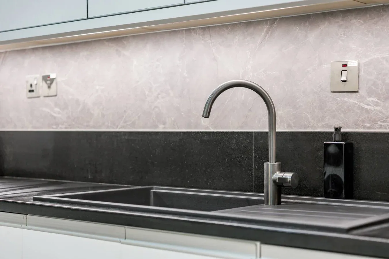 A kitchen sink with a curved faucet sits beneath a dark backsplash.