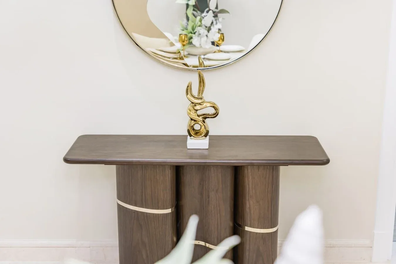 A console table beneath a round mirror, with a gold-colored decorative sculpture.