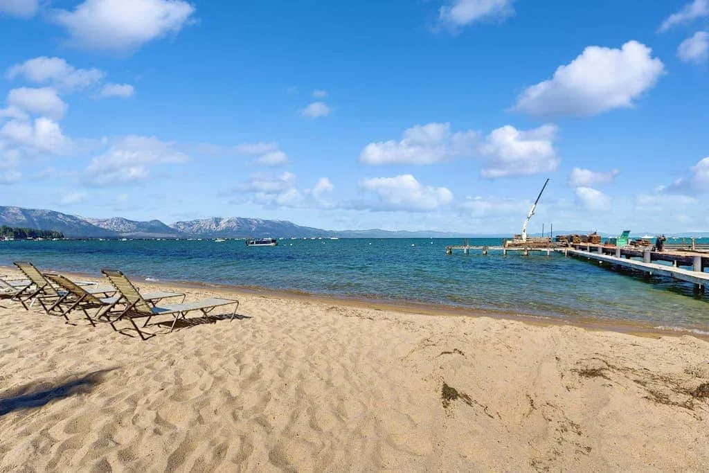 Una playa de arena junto a aguas azules tranquilas, con tumbonas y un muelle.