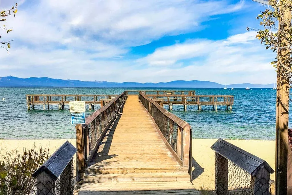 Un muelle de madera se extiende desde una playa de arena hacia aguas azules y colinas lejanas.