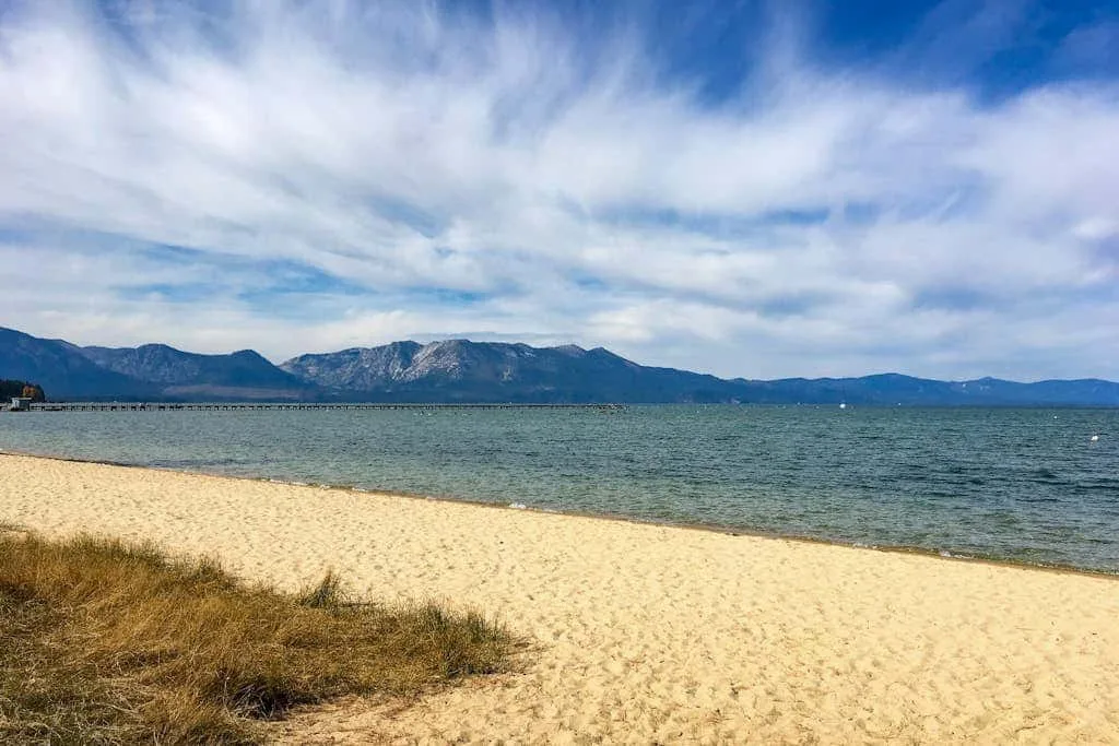 Una orilla de arena se extiende junto al agua tranquila, con montañas a lo lejos.