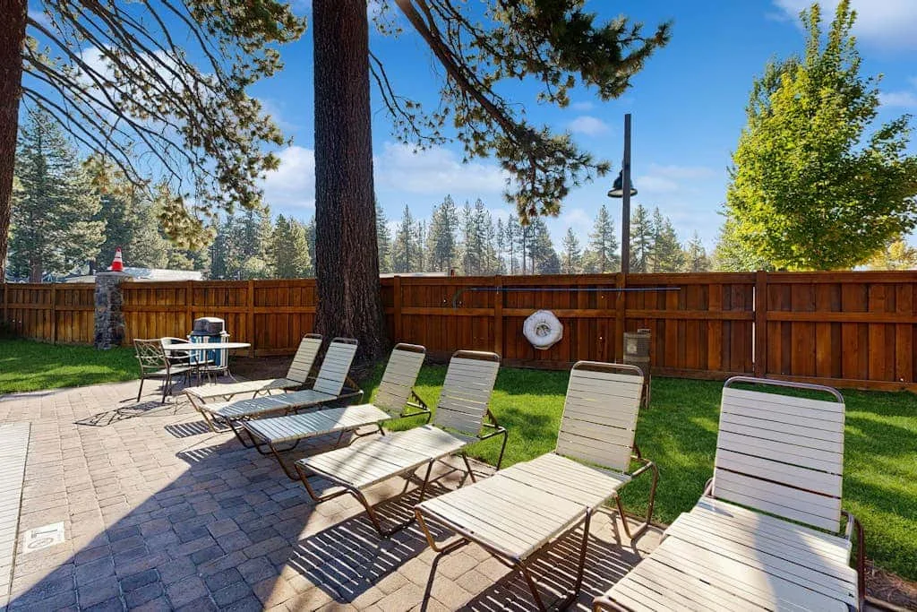 Un patio exterior cercado con asientos reclinables y una zona pavimentada bajo los árboles.